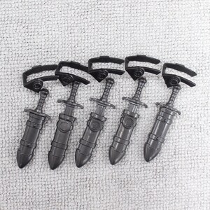 May include: Six black plastic toy swords with sheaths. The swords have a simple design with a cross guard and a pointed tip. The sheaths are decorated with a single band.