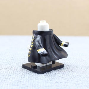May include: Black and gold Lego minifigure wearing a long black coat with gold buttons. The minifigure is standing on a black base.