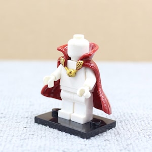 May include: A white Lego minifigure wearing a red cape with a gold collar and a gold amulet. The minifigure is standing on a black base.