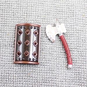 May include: A miniature shield and axe set. The shield is bronze-colored with a decorative border and diamond-shaped accents. The axe has a silver head and a red handle. The set is designed for a miniature figure or display.