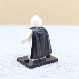 May include: A white Lego figure with a black cape, standing on a black base. The figure has a white head and arms. The cape drapes over the shoulders and back, adding a dramatic flair to the miniature character.
