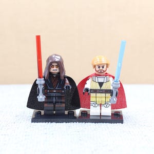 May include: Two Lego minifigures, one with a red lightsaber and dark clothing, the other with a blue lightsaber and light-colored attire. Both figures have capes and stand on black bases.