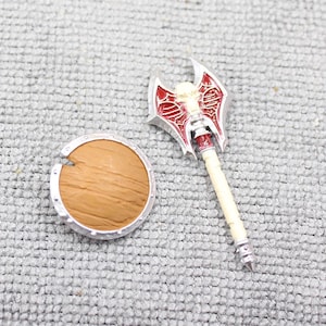 May include: A miniature toy axe with a red and silver blade and a cream-colored handle, next to a small wooden shield with a silver rim. The axe features a skull design.