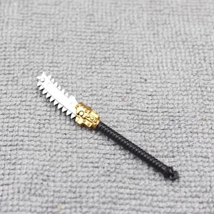 May include: A miniature silver and gold chainsaw with a black handle. The saw blade has sharp teeth, and the handle is textured. The gold section has intricate details. This toy weapon is likely for a miniature figure or model.