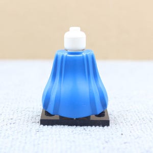 May include: A blue plastic LEGO minifigure skirt with a white LEGO minifigure head on top. The skirt is on a black LEGO baseplate.