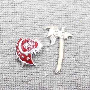 May include: A silver and red miniature shield and axe set. The shield is red with silver trim and a central star design. The axe has a silver head and a cream-colored handle. The set is designed for fantasy or role-playing games.