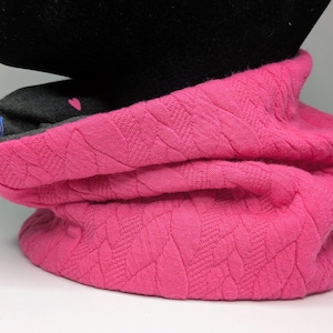 Könnte beinhalten: Ein leuchtend rosa Nackenwärmer mit einem Steppmuster. Die Innenseite ist grau mit einem kleinen rosa Herz und einem bunten Streifendesign. Der Nackenwärmer wird auf einem schwarzen Schaufensterkopf präsentiert.