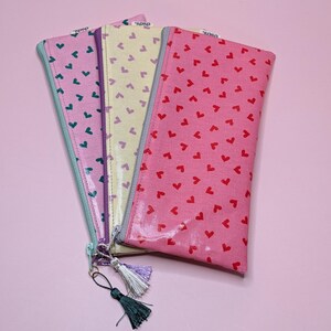 Könnte beinhalten: Drei Reißverschlusstaschen mit Herzmustern. Die Taschen sind rosa, gelb und lila mit roten, grünen und blauen Herzen. Jede Tasche hat einen Quasten.