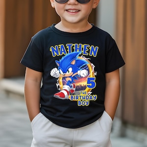 K&ouml;nnte beinhalten: Schwarzes T-Shirt mit einer Grafik von Sonic the Hedgehog, dem Namen "NATHEM" dar&uuml;ber und "5 THE BIRTHDAY BOY" darunter. Das Design beinhaltet eine Stadtlandschaft und einen Ring. Das T-Shirt wird von einem Kind getragen.
