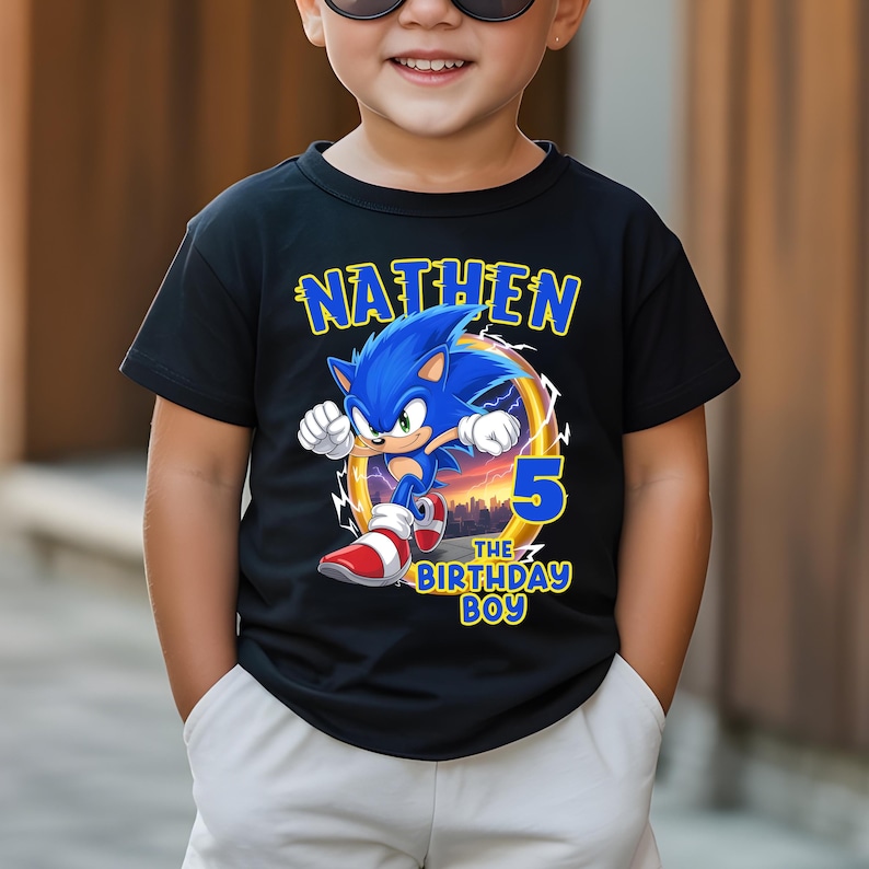 K&ouml;nnte beinhalten: Schwarzes T-Shirt mit einer Grafik von Sonic the Hedgehog, dem Namen "NATHEM" dar&uuml;ber und "5 THE BIRTHDAY BOY" darunter. Das Design beinhaltet eine Stadtlandschaft und einen Ring. Das T-Shirt wird von einem Kind getragen.