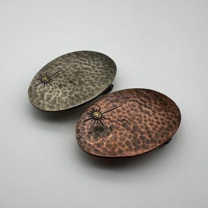 Può includere: Due fibbie ovali per cintura in metallo con una texture martellata. Una fibbia è argentata con un design a stella dorata, l'altra è in rame con un design a stella dorata.