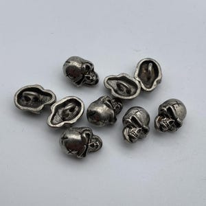 May include: Eight silver skull-shaped buttons with a hole for sewing on. The buttons are scattered on a white surface.