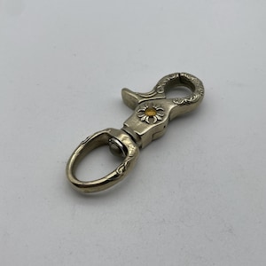Puede incluir: Un llavero de metal plateado con un diseño adornado y un pequeño acento de flor amarilla.