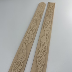 May include: Two strips of light brown leather with a decorative pattern of feathers carved into the surface.