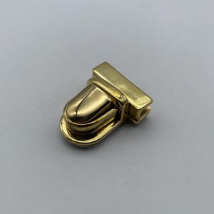 Pode incluir: Um fecho de metal dourado com uma parte superior arredondada ligeiramente curva e uma base retangular. O fecho é projetado para ser usado para prender correias ou cintos.