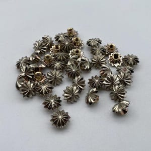 Op de afbeelding: Een stapel zilverkleurige metalen stervormige studs met een verhoogd midden. De studs hebben een decoratief, geribbeld ontwerp rond de randen.