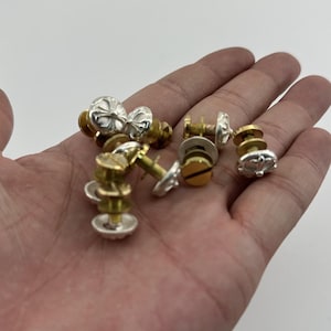 May include: A collection of silver and gold-toned metal screw-back earrings. The earrings have decorative tops with various designs, including floral and geometric patterns. The earrings are displayed against a neutral background.