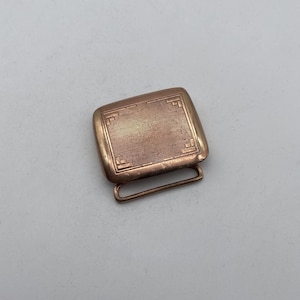 Puede incluir: Una hebilla de cinturón de metal dorado con un diseño rectangular y un patrón geométrico.