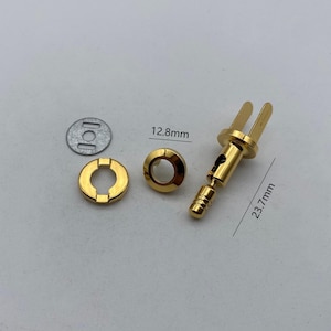 Puede incluir: Un conjunto de conectores y componentes eléctricos de color dorado. El conector principal mide 23,7 mm de largo, con un anillo de 12,8 mm de diámetro y una pequeña pieza metálica circular. Los componentes están dispuestos sobre un fondo claro.