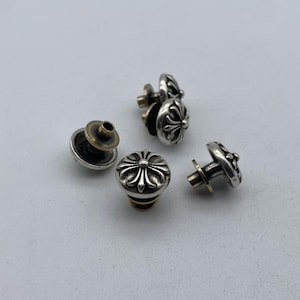 May include: A collection of silver-colored metal studs with a decorative cross design. The studs have a detailed, ornate appearance and are set against a plain, light background. The metal studs have a brass-colored post.