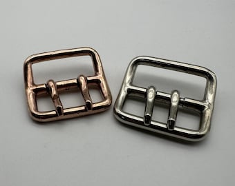 May include: Two metal belt buckles, one is copper and the other is silver. Both buckles have a rectangular shape with three prongs.