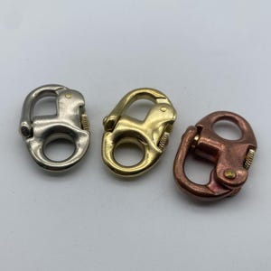 Könnte beinhalten: Drei Metallschließen in Silber-, Gold- und Kupferausführung. Jede Schließe hat eine drehbare Öse und einen federbelasteten Verschluss.