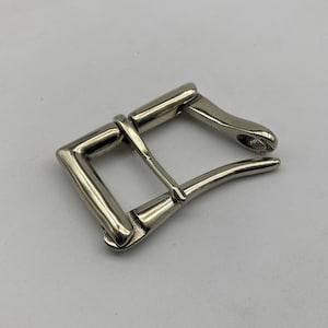 Pode incluir: Fivela de cinto prateada com estrutura retangular e lingueta curva. A fivela é feita de metal e tem um acabamento brilhante. O design é simples e clássico, adequado para vários estilos de cinto.