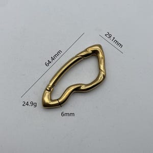 Pode incluir: Um mosquetão de metal dourado com um design ondulado único. Mede 6,4 cm de comprimento, 2,9 cm de largura e 0,6 cm de espessura. Pesa 24,9 gramas.