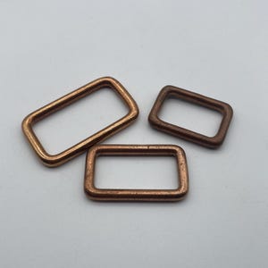 May include: Three rectangular metal rings in different sizes. The rings are a copper color and have a smooth, polished finish.