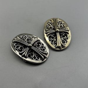 May include: Two oval-shaped silver-toned metal buttons or decorative accents. Each piece features an intricate, openwork filigree pattern surrounding a central raised cross design with flared arms and a small circular stud at the intersection point.