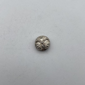 May include: A small, silver-colored, domed button with a four-petal floral design. The button has a slightly textured surface and is set against a plain, light gray background. The button is likely made of metal.