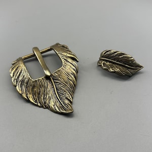 Può includere: Un set di due pezzi di ferramenta per cintura in metallo color ottone antico. Uno è una fibbia per cintura a forma di piuma, e l'altro è un piccolo pezzo a forma di piuma. La fibbia ha un telaio rettangolare e un perno.