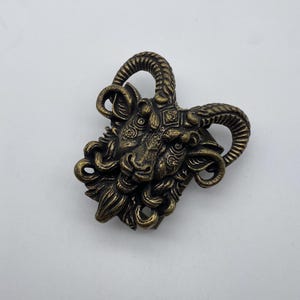 Puede incluir: Una hebilla de cinturón de metal dorado detallada y adornada con una cabeza de carnero estilizada con cuernos.