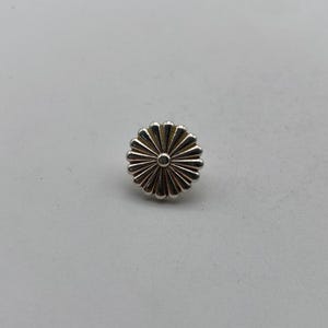 May include: A silver-toned metal pin with a flower design. The flower has a central circle and radiating petals.
