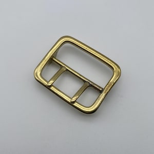 May include: A rectangular brass buckle with three bars. The buckle has rounded corners and a slightly aged appearance. The metal has a warm, golden hue. This hardware component is suitable for belts, bags, or other leathercraft projects.