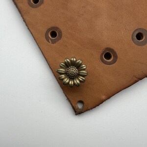 May include: A close-up of a brass daisy-shaped decorative nail on a piece of brown leather.