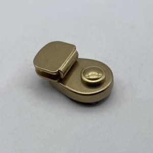 Pode incluir: Um fecho de metal dourado com um botão redondo. O fecho é projetado para ser usado para prender correias ou outros materiais.