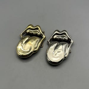 May include: Two belt buckles shaped like a mouth with a protruding tongue. One buckle is finished in a polished gold tone, while the other is finished in a polished silver tone. Both items feature a smooth, reflective metallic surface.