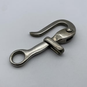 Puede incluir: Un mosquetón de metal plateado con un extremo de gancho curvado y un extremo de anillo. El gancho tiene un mecanismo de bisagra para un cierre seguro. El metal tiene un aspecto ligeramente desgastado.