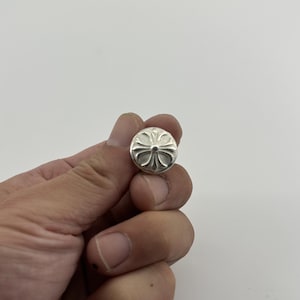 Puede incluir: Un pequeño botón redondo plateado con un diseño en forma de cruz. El botón se sostiene en una mano sobre un fondo blanco. El diseño del botón presenta un elemento central con cuatro brazos curvos que se extienden hacia afuera.