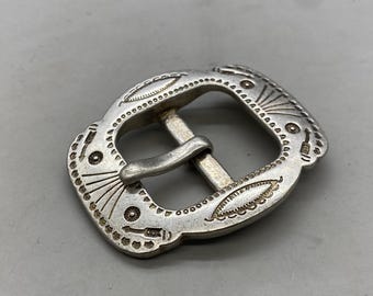 May include: Silver belt buckle with an intricate design. The buckle is rectangular with rounded corners and features a detailed pattern of lines and dots.