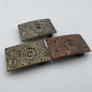May include: Three rectangular metal belt buckles in bronze, brass, and copper tones. Each buckle features a textured surface with a central circular design and smaller surrounding circles. The buckles are displayed on a white background.