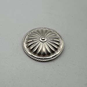 Op de afbeelding: Een zilverkleurig, rond decoratief object met een bloemmotief. Het midden heeft een klein gaatje en de buitenrand heeft een touwachtige textuur. Het object is waarschijnlijk van metaal en heeft een glanzende afwerking.