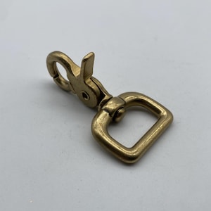 Könnte beinhalten: Ein goldfarbener, drehbarer Haken aus Metall mit einem D-Ring. Der Haken hat einen federbelasteten Verschluss.