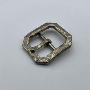 Puede incluir: Hebilla geométrica plateada con superficie texturizada. La hebilla tiene forma rectangular con bordes biselados y una barra y pasador centrales para asegurar un cinturón o correa. El metal tiene un aspecto ligeramente envejecido.