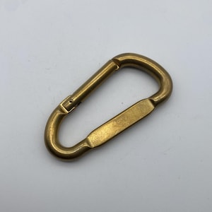 May include: A gold-colored metal carabiner clip. The clip is closed and has a smooth, shiny finish.