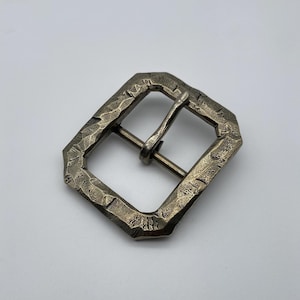 Könnte beinhalten: Eine silberfarbene Gürtelschnalle aus Metall mit einer strukturierten, gehämmerten Oberfläche. Die Schnalle hat eine rechteckige Form mit abgerundeten Ecken und einem leicht erhöhten Rand.