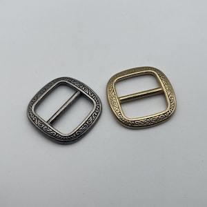 May include: Two ornate metal buckles, one silver and one gold, with a square shape and intricate floral designs around the edges.
