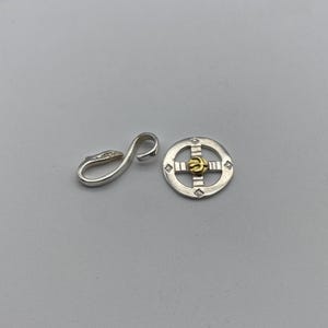 May include: A silver and gold pendant with a circular design. The pendant has a cross shape with a small gold circle in the center. The pendant is attached to a silver clasp.
