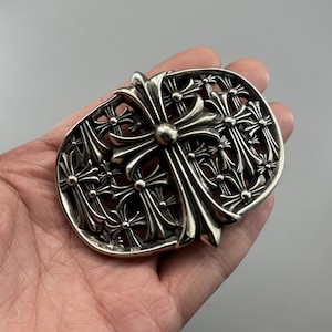 May include: A polished silver belt buckle featuring an intricate openwork design composed of multiple layered cross motifs. The central cross is larger and more prominent, surrounded by smaller, repeating cross shapes within an oval frame.